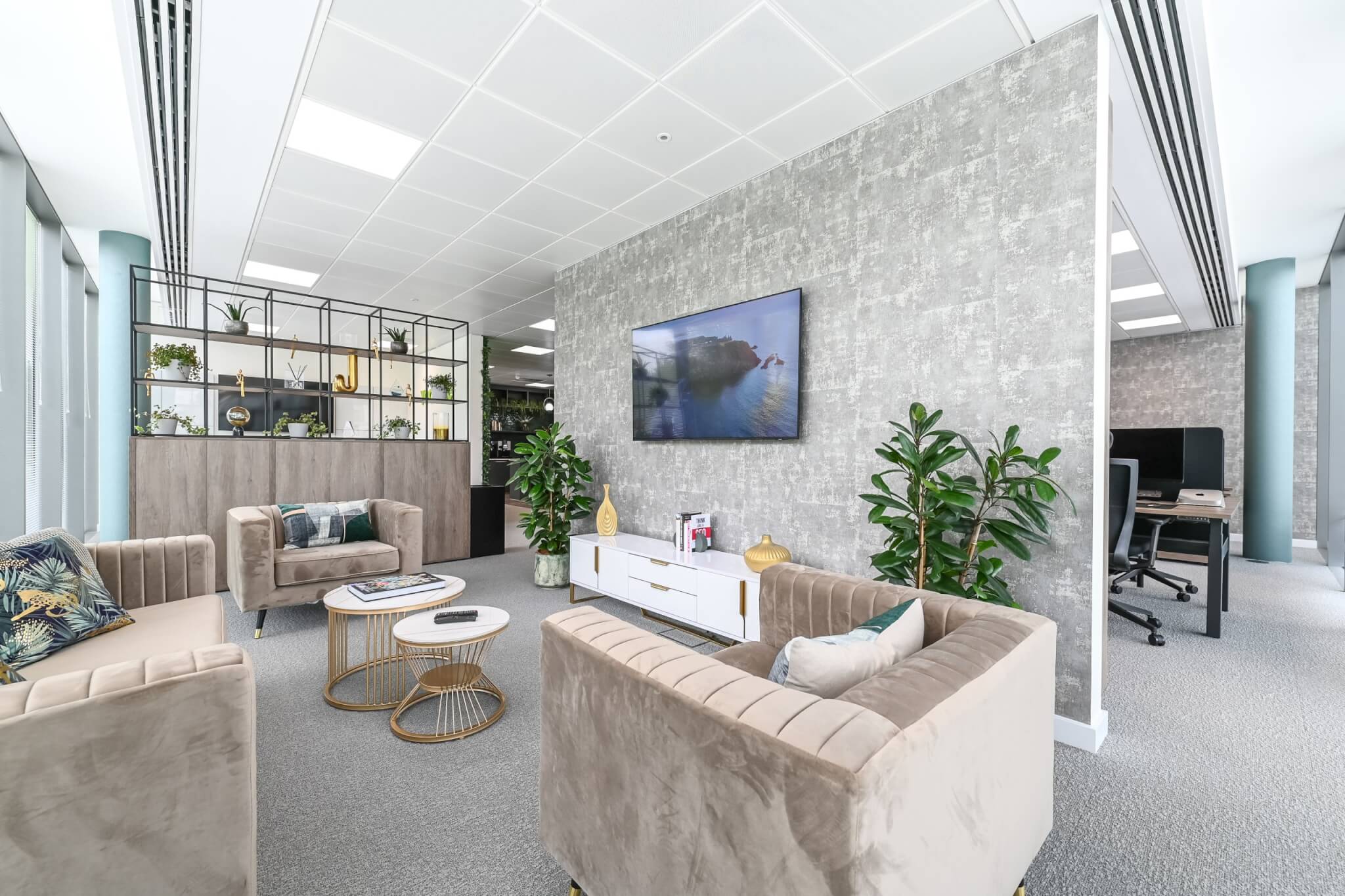 Lounge seating area with a wall-mounted TV, plants and a breakout desk beyond