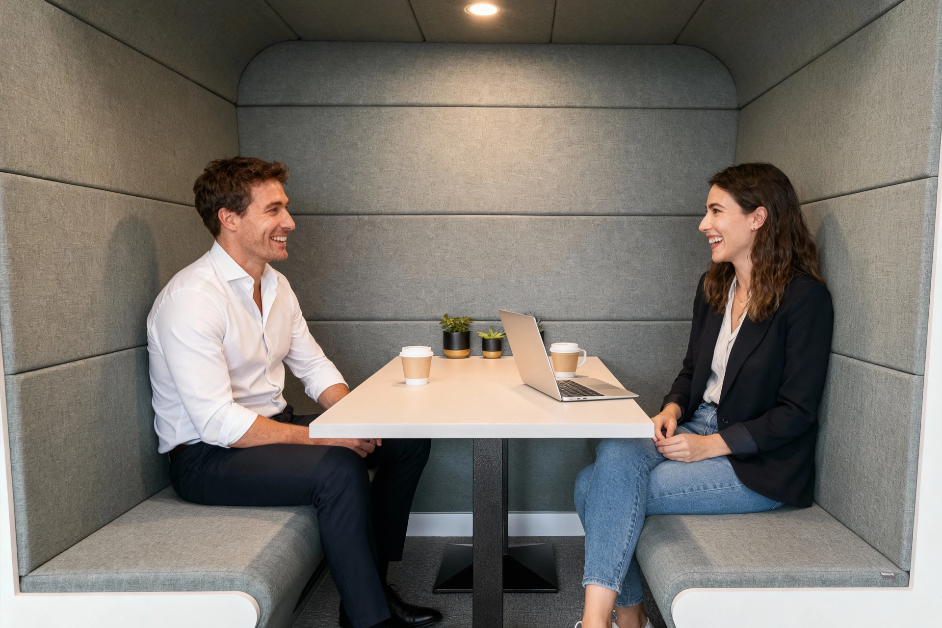 Two colleagues laughing together over coffee in a private meeting pod
