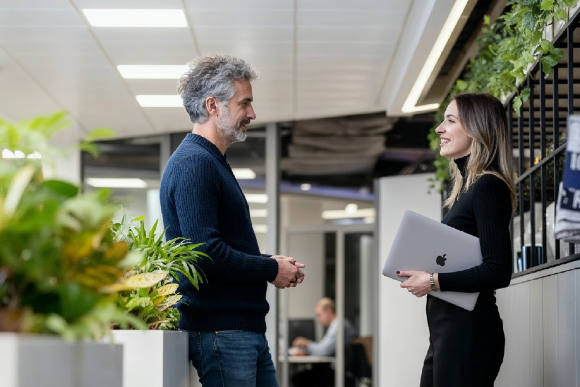 Two colleagues chatting in an office corridor, one holding a laptop